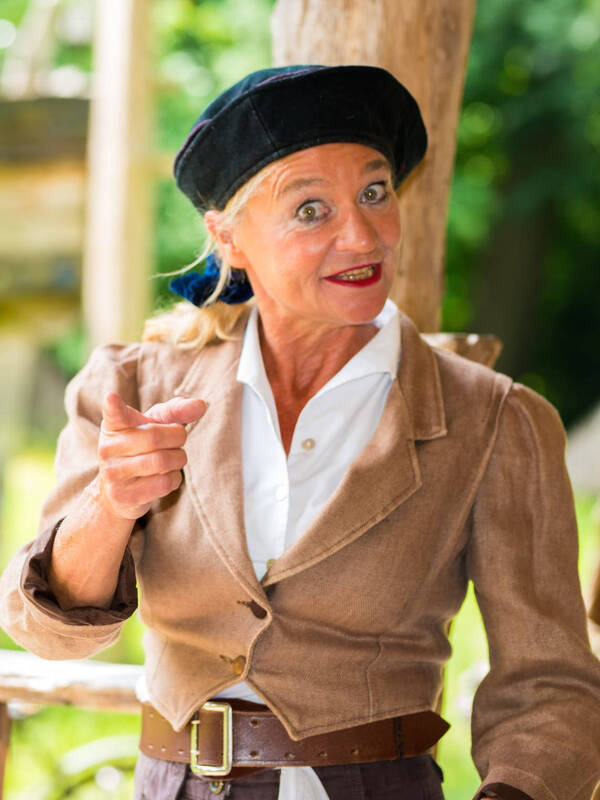 Schauspielerin Karin Schroeder blickt und zeigt in die Kamera, im Hintergrund sind grüne Bäume und Büsche zu sehen.