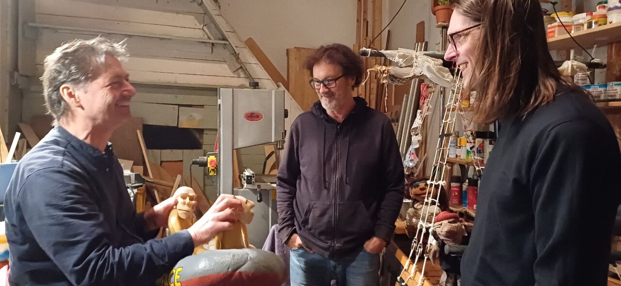 Christoph Buchfink lacht und zeigt eine Holzpuppe. Karl-Heinz Ahlers schaut interessiert zu. Mark Roberts lacht. Drum herum ist Buchfinks Werkstatt zu sehen: Farbtöpfe, Material, ein kleines Puppen-Boot.