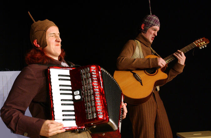 B&uuml;hnenfoto: Herr Pling mit Gitarre rechts und Frau Plong mit Akkordeon links machen Musik und kneifen dabei die Augen zu und verziehen das Gesicht als w&auml;re es sehr laut.