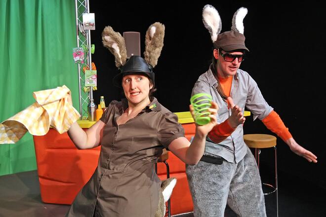 B&uuml;hnenfoto: Im Vordergrund links h&auml;lt Bardame Hasi in ihrer rechten Hand ein wei&szlig;-gelb-kariertes Geschirrtuch und in ihrer linken Hand ein gr&uuml;n gestreiftes Glas. Rechts neben ihr gestikuliert DJ Hop Hop.