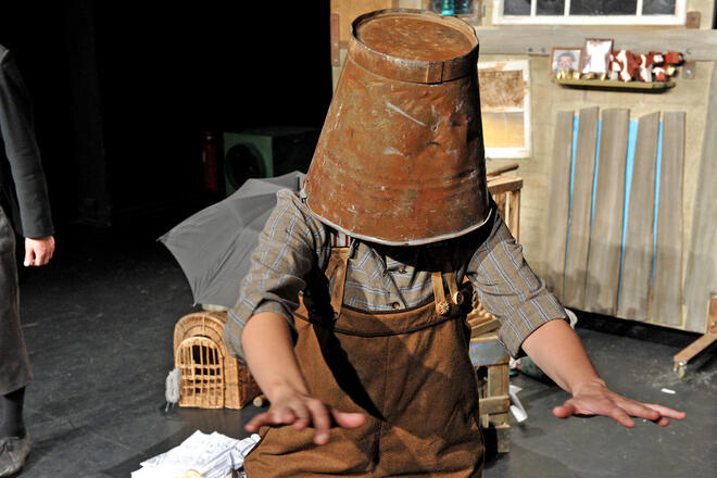 B&uuml;hnenfoto: Im Vordergrund Mama Muh mit einem Eimer auf dem Kopf und nach vorne ausgestreckten Armen.