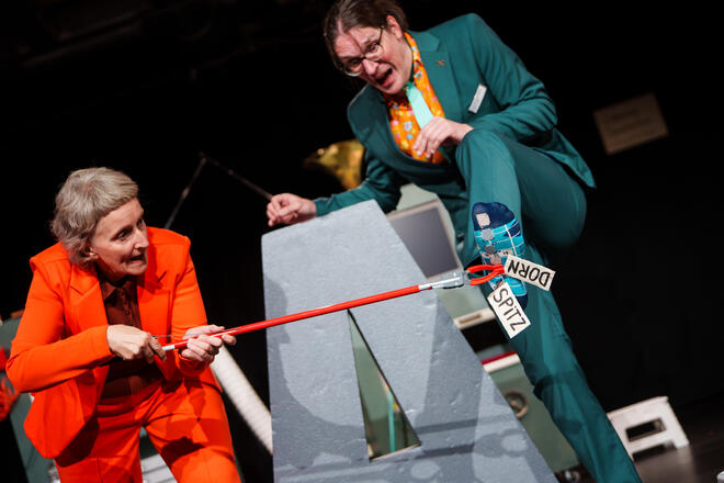 B&uuml;hnenfoto: Dr. Patsch h&auml;lt seinen Fu&szlig; hoch. Daran kleben sichtbar kleine W&ouml;rter wie "spitz" und "Dorn". Professorin Toll versucht diese konzentriert mit einem gro&szlig;en roten Greifer zu entfernen. Dr. Patsch macht ein &auml;ngstlich verzerrtes Gesicht.